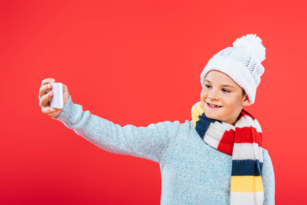 χαμογελαστός παιδί σε καπέλο και κασκόλ παίρνοντας selfie απομονώνονται σε κόκκινο - Φωτογραφία, εικόνα