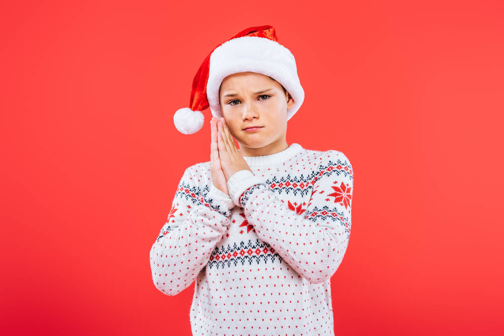 sad kid in sweater and santa hat showing please gesture isolated on red - Photo, Image