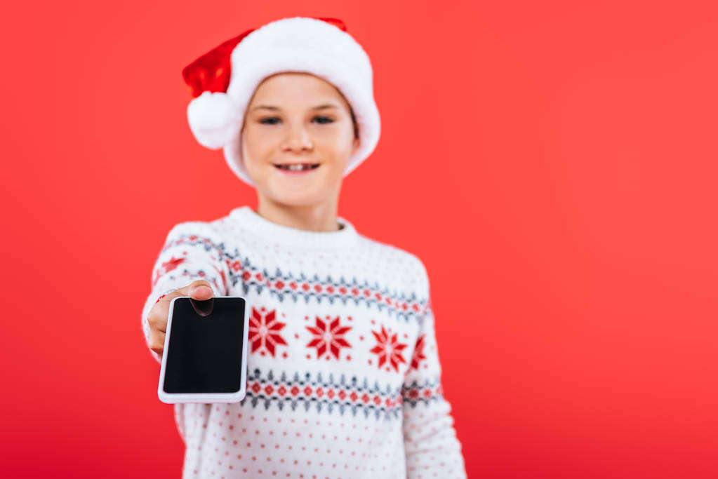 foco selectivo del niño sonriente en el teléfono inteligente de la celebración del sombrero de santa con la pantalla en blanco aislada en rojo
 - Foto, imagen