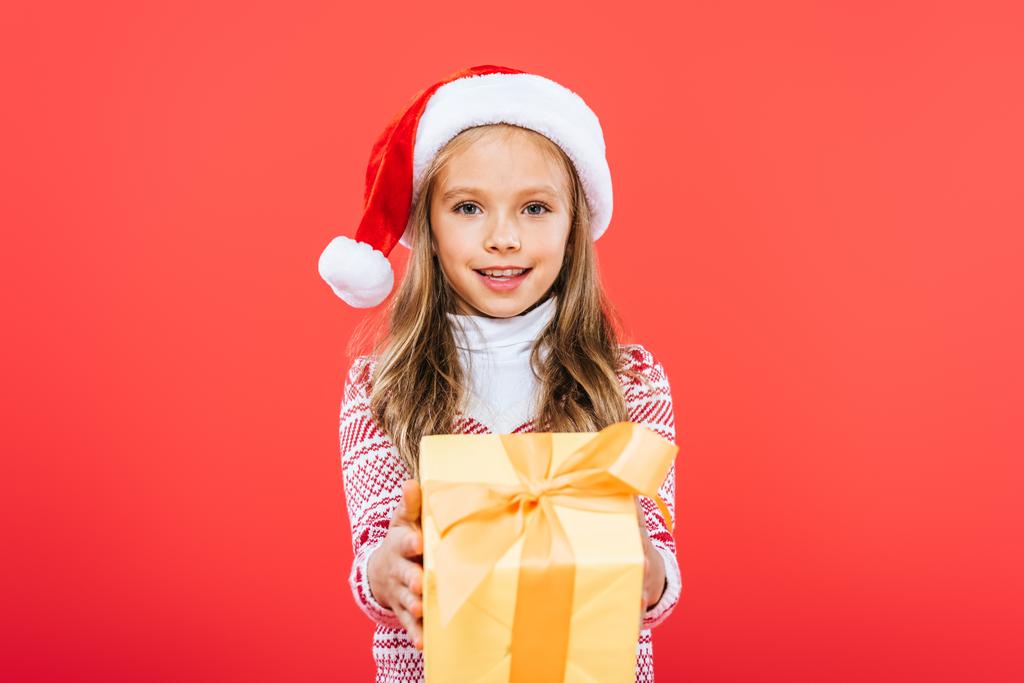 front view of smiling kid in santa hat holding present isolated on red - Photo, Image