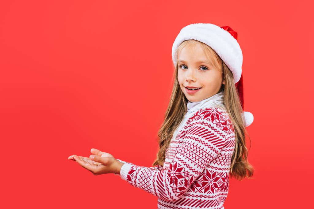 mosolygós gyerek a pulóvert és Santa kalap a kinyújtott kezet izolált vörös - Fotó, kép
