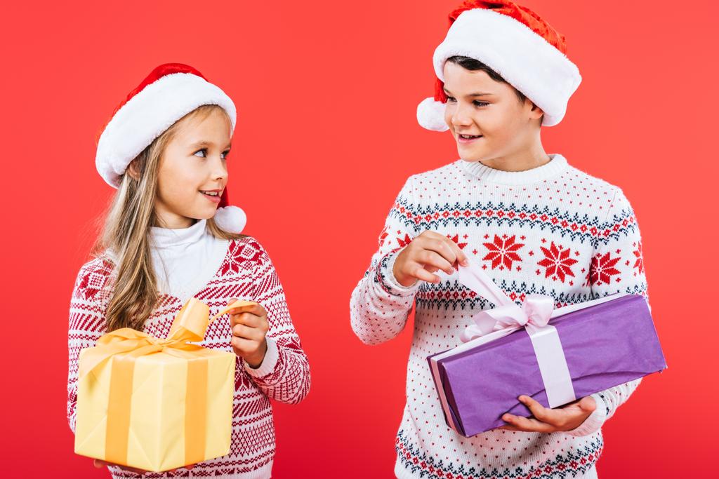 duas crianças sorridentes em chapéus de Papai Noel com presentes olhando um para o outro isolado no vermelho
 - Foto, Imagem