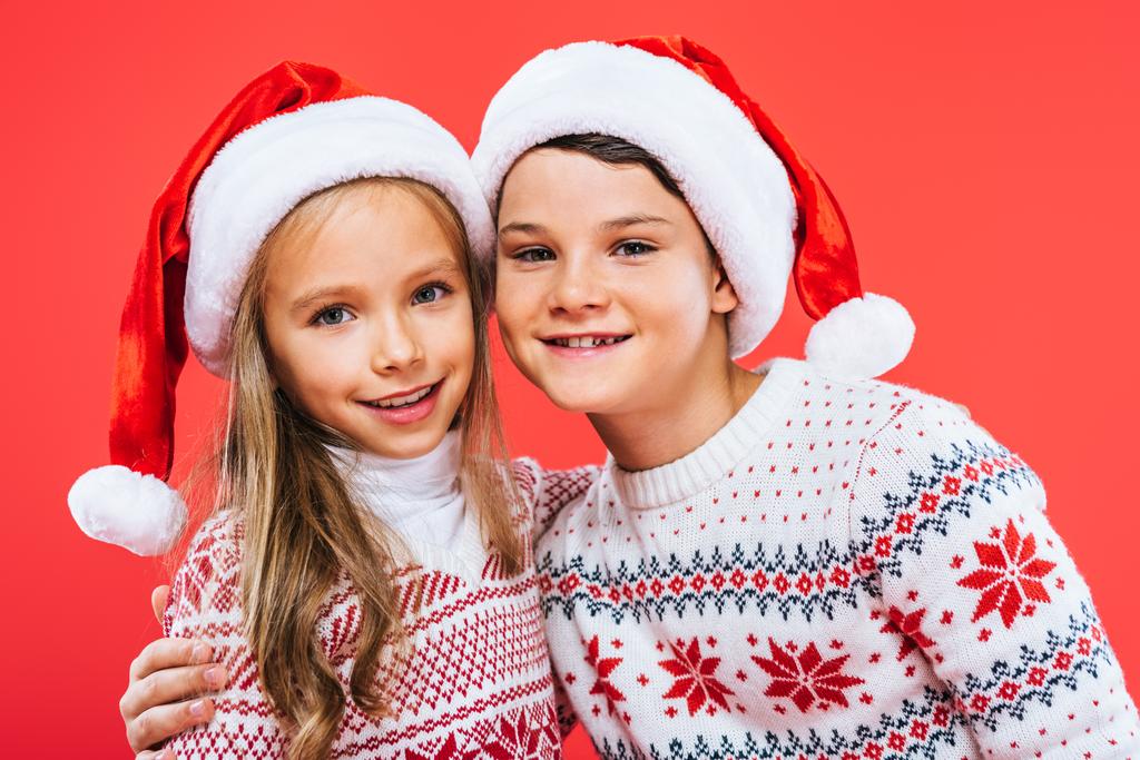 Frontansicht zweier lächelnder Kinder in Weihnachtsmannmützen und Pullovern, die sich vereinzelt auf Rot umarmen - Foto, Bild