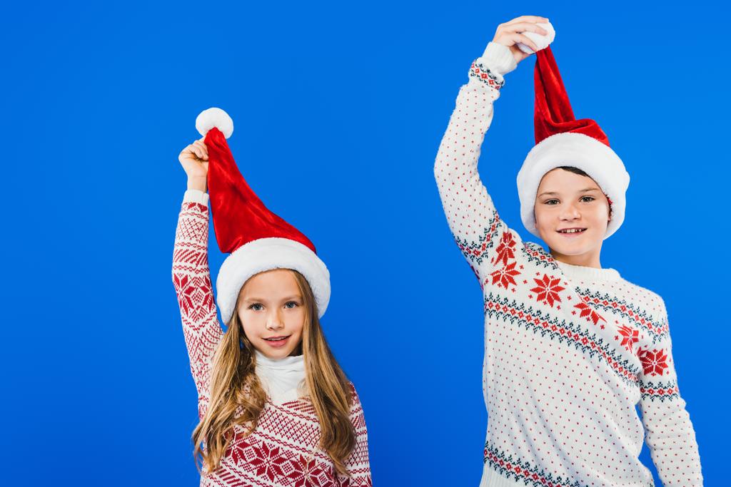 vista frontal de dos niños sonrientes en suéteres y sombreros de santa aislados en azul
 - Foto, Imagen