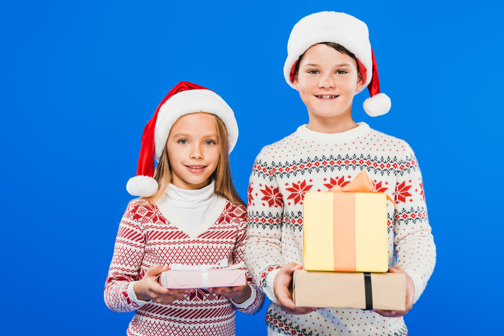 vista frontal de dos niños sonrientes en sombreros de santa con regalos aislados en azul
 - Foto, imagen