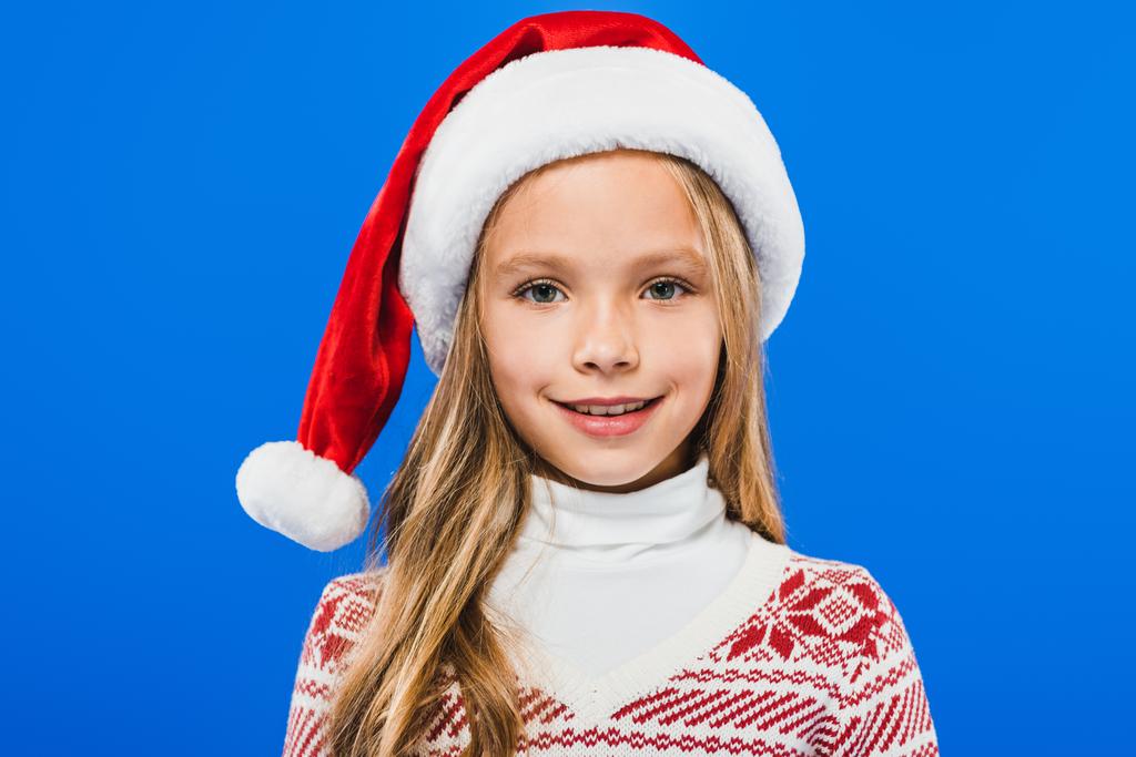 pohled na usmívající se dítě v Santa klobouku a svetru izolovaně na modrém - Fotografie, Obrázek