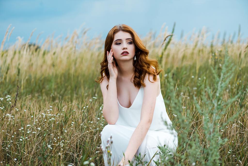 フィールドに座っている白いドレスを着た若い赤毛の女性の選択的な焦点  - 写真・画像