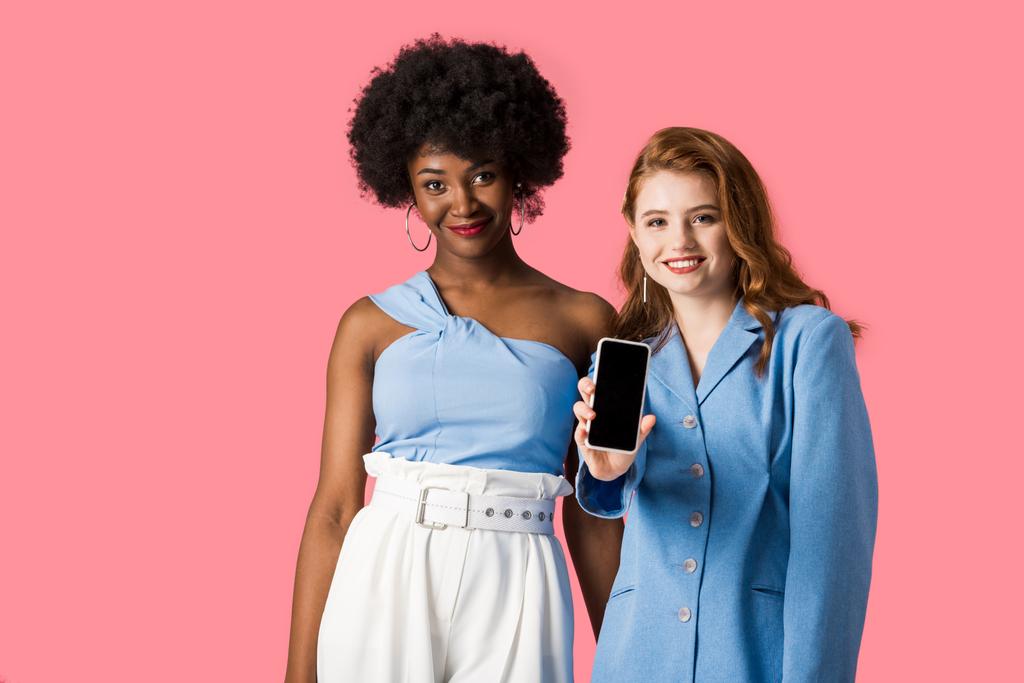 ピンクで孤立したアフリカ系アメリカ人女性の近くに空白の画面を持つスマートフォンを持つ幸せな赤毛の女の子  - 写真・画像
