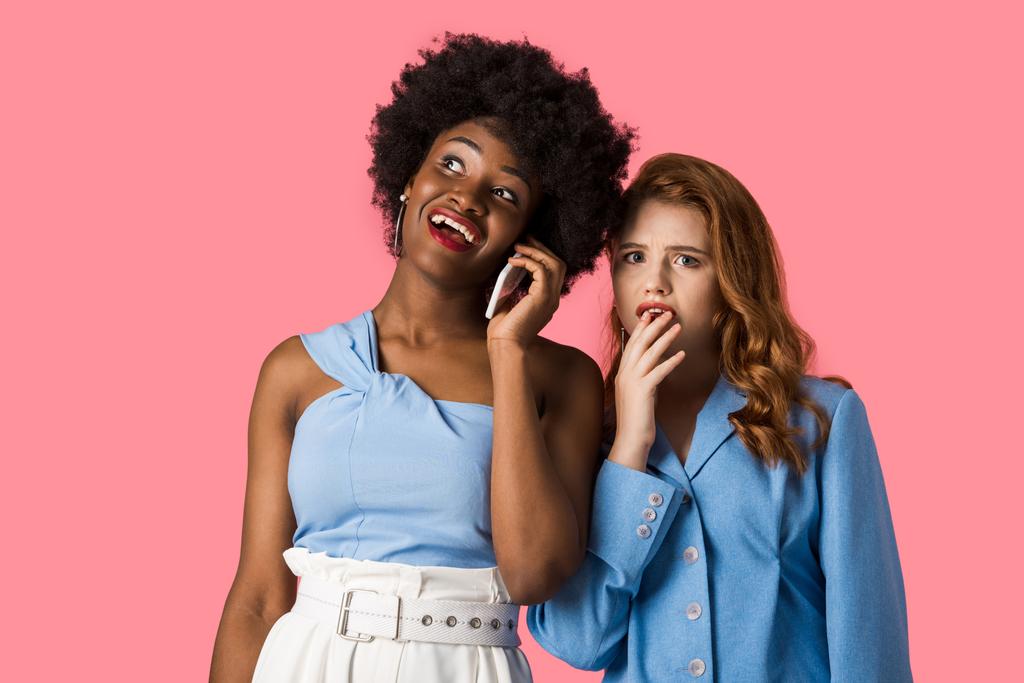šťastná Africká Američanka mluvila o smartphone nedaleko ustaraně červené dívky izolované na růžovém  - Fotografie, Obrázek