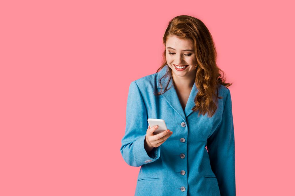 chica pelirroja sonriente usando teléfono inteligente aislado en rosa
  - Foto, imagen