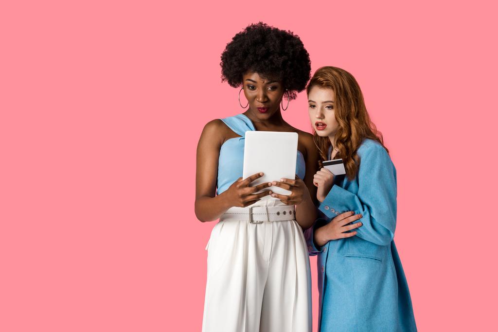 chica pelirroja bastante celebración de la tarjeta de crédito cerca de la mujer afroamericana con tableta digital aislado en rosa
  - Foto, Imagen