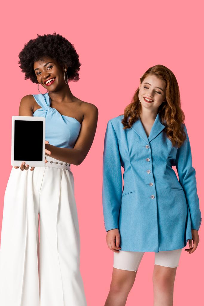 wesoły rudowłosy dziewczyna patrząc na African American kobieta gospodarstwa cyfrowego tabletka z pustym ekranie na białym tle różowy - Zdjęcie, obraz
