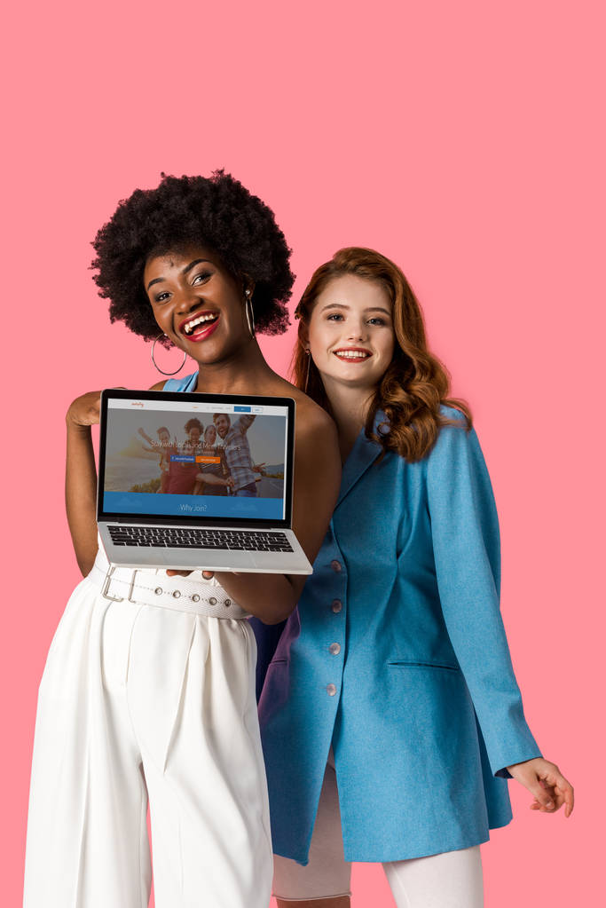 KYIV, UKRAINE - AUGUST 9, 2019: cheerful multicultural girls smiling near laptop with couchsurfing website on screen isolated on pink  - Photo, Image
