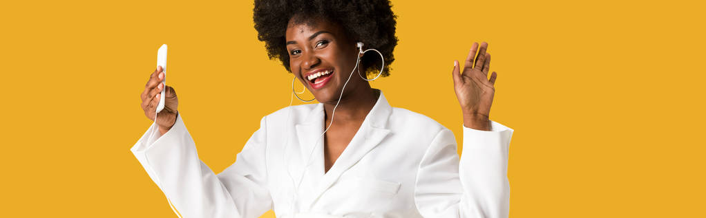 tiro panorâmico de menina americana africana alegre ouvindo música enquanto segurando smartphone e gesticulando isolado em laranja
  - Foto, Imagem