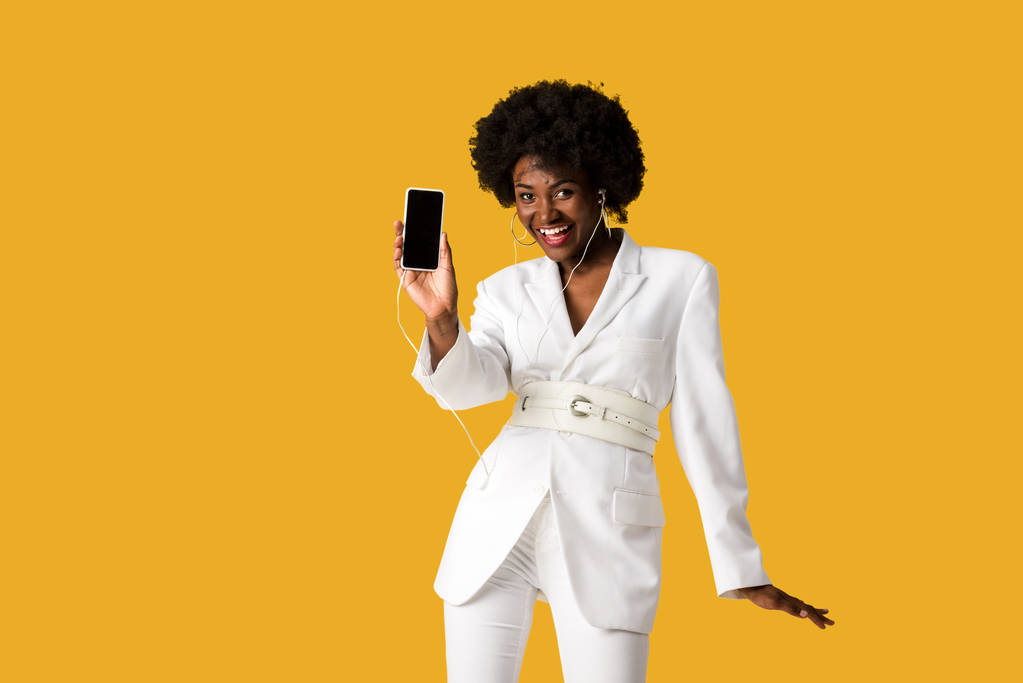 オレンジ色に隔離された空白の画面でスマートフォンを保持しながら、音楽を聴いているアフリカ系アメリカ人の女の子を笑顔  - 写真・画像