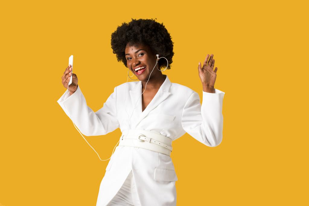 улыбающаяся африканская американская девушка слушает музыку, держа в руках смартфон и жестикулируя изолированным на оранжевом
  - Фото, изображение
