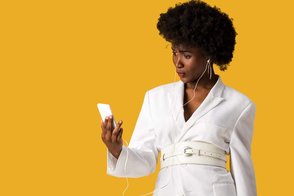 удивленный африканский американец женщина держит смартфон и слушать музыку изолированы на оранжевый
 - Фото, изображение