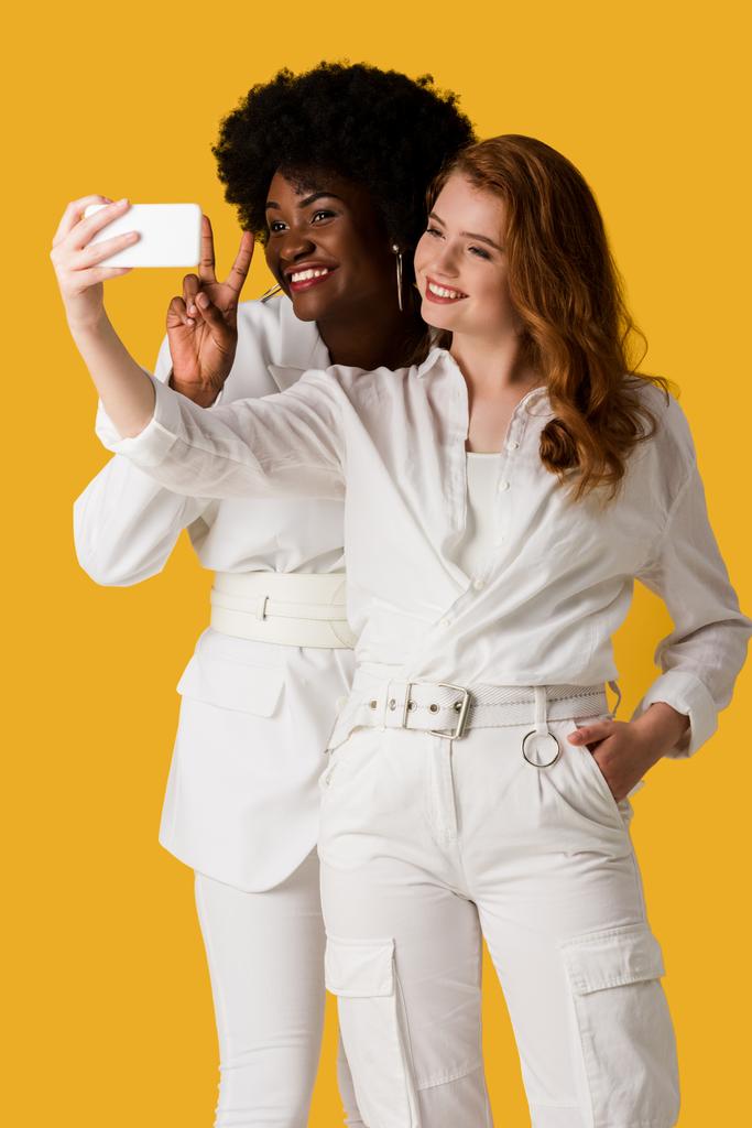 χαμογελαστά πολυπολιτισμικά κορίτσια παίρνοντας selfie απομονωμένη στο πορτοκαλί  - Φωτογραφία, εικόνα