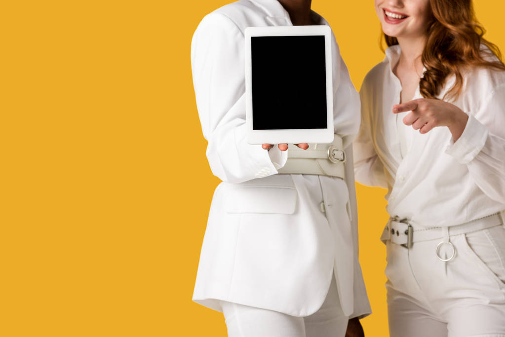 abgeschnittene Ansicht einer afrikanisch-amerikanischen Frau, die ein digitales Tablet mit leerem Bildschirm in der Nähe eines glücklichen rothaarigen Mädchens hält, das auf orange isoliert ist  - Foto, Bild