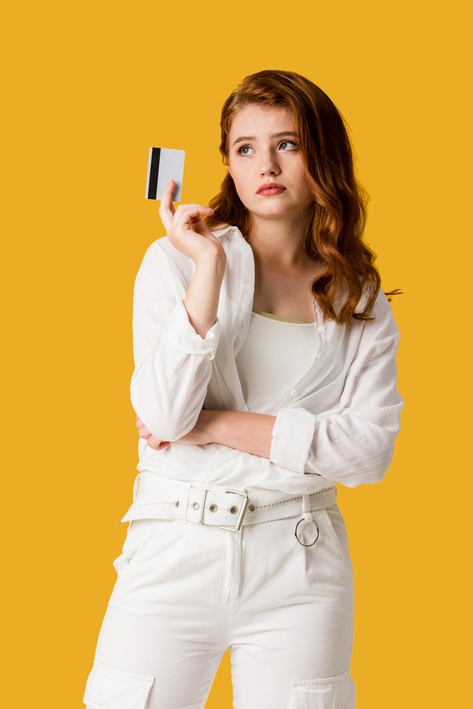 σκεπτική κορίτσι κρατώντας πιστωτική κάρτα απομονωμένη σε πορτοκαλί  - Φωτογραφία, εικόνα