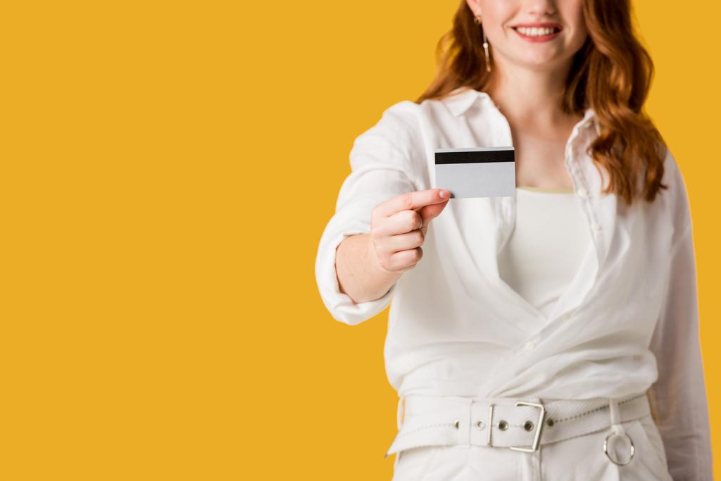 περικομμένη όψη του χαρούμενο κοκκινομάλλα κορίτσι κρατώντας πιστωτική κάρτα απομονώνεται σε πορτοκαλί  - Φωτογραφία, εικόνα