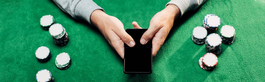 ポーカーチップの近くに空白の画面でスマートフォンを保持している男のパノラマショット - 写真・画像