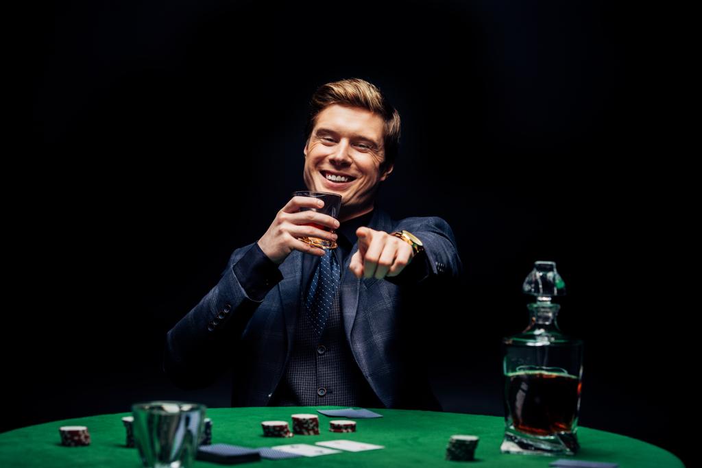 黒で隔離されたウイスキーでガラスを保持しながら、指で指を指差す幸せな男の選択的な焦点  - 写真・画像