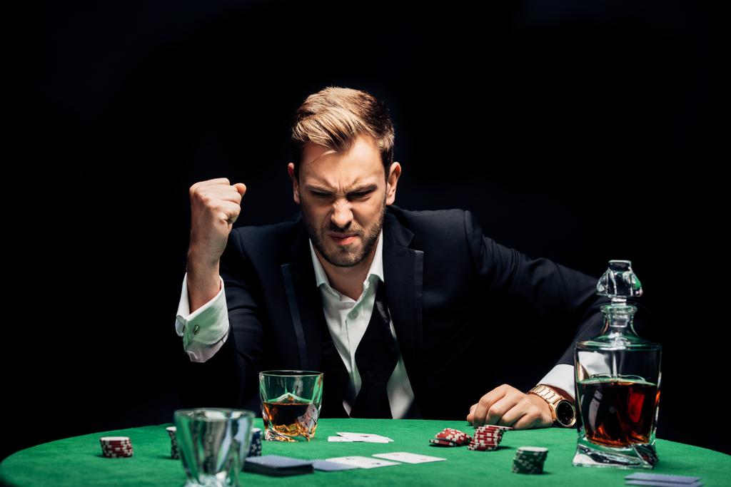 selektywne skupienie wściekłego człowieka z zaciśniętej ręki blisko alkoholu i karty do gry odizolowane na czarno  - Zdjęcie, obraz