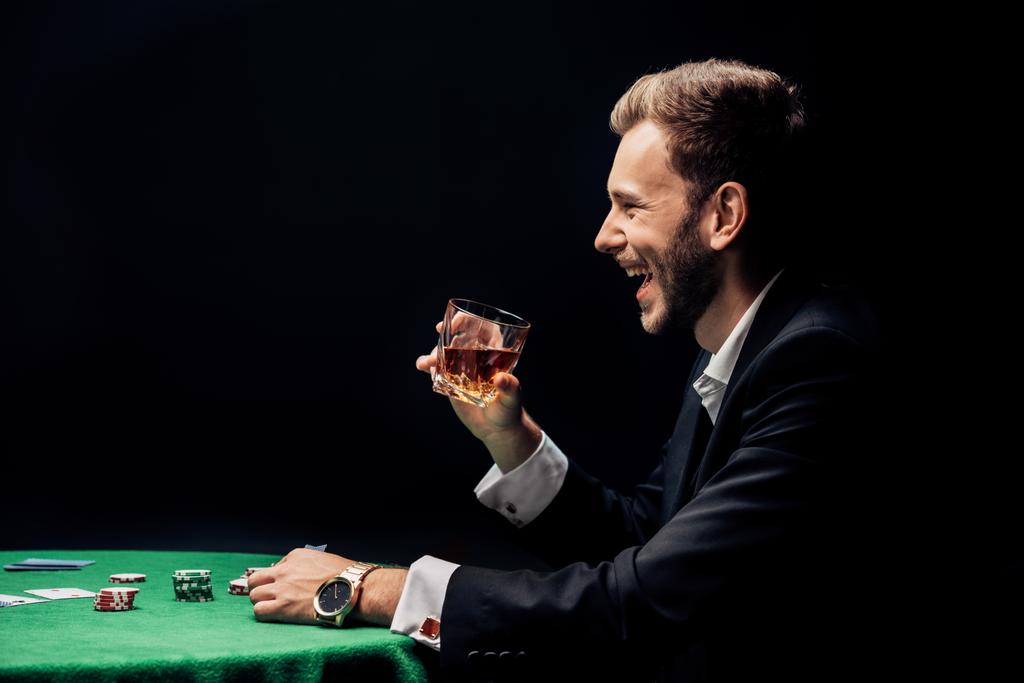 Widok z boku szczęśliwego Brodatego człowieka uśmiechając się i trzymając szkło w pobliżu stołu pokerowego na czarno  - Zdjęcie, obraz