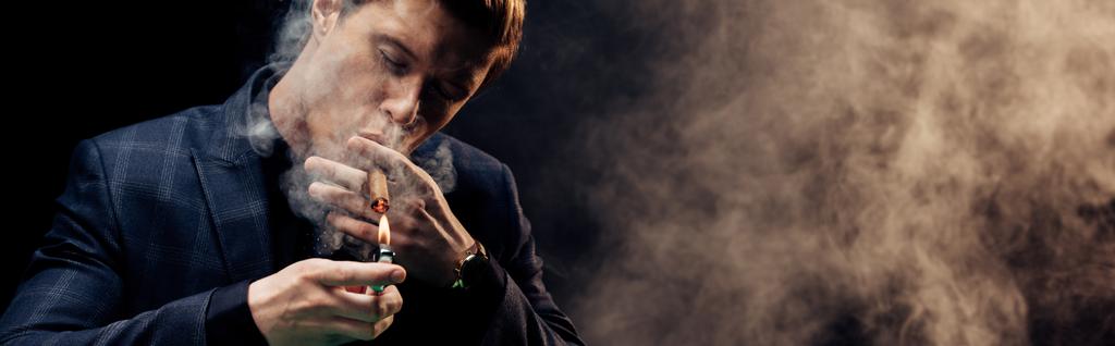 Panoramaaufnahme eines gutaussehenden Mannes mit Feuerzeug beim Rauchen auf schwarz mit Rauch  - Foto, Bild