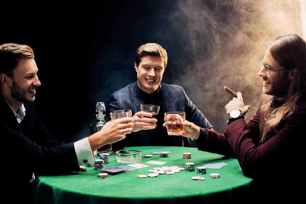 KYIV, UKRAINE - 20 de agosto de 2019: homens felizes segurando óculos com álcool perto da mesa de poker em preto com fumaça
 - Foto, Imagem