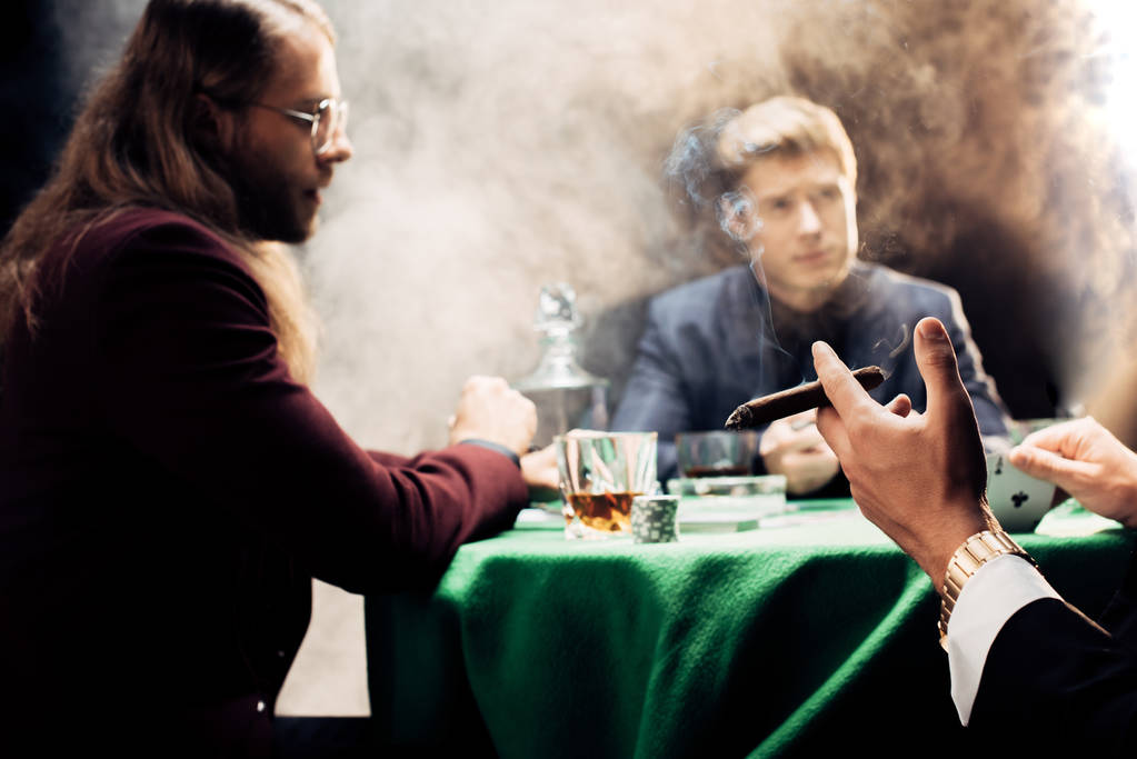 bijgesneden weergave van de man die sigaar houdt tijdens het spelen van poker op zwart met rook  - Foto, afbeelding