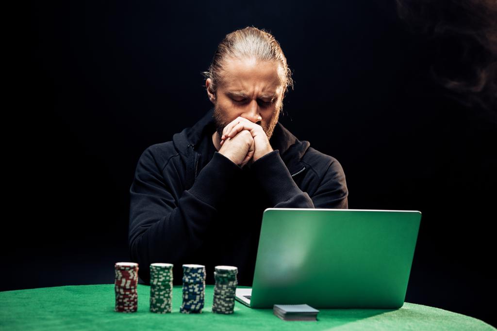 KYIV, UCRAINA - AGOSTO 20, 2019: uomo che copre la faccia durante l'utilizzo di laptop vicino a chip di poker e carte da gioco su nero con fumo
 - Foto, immagini