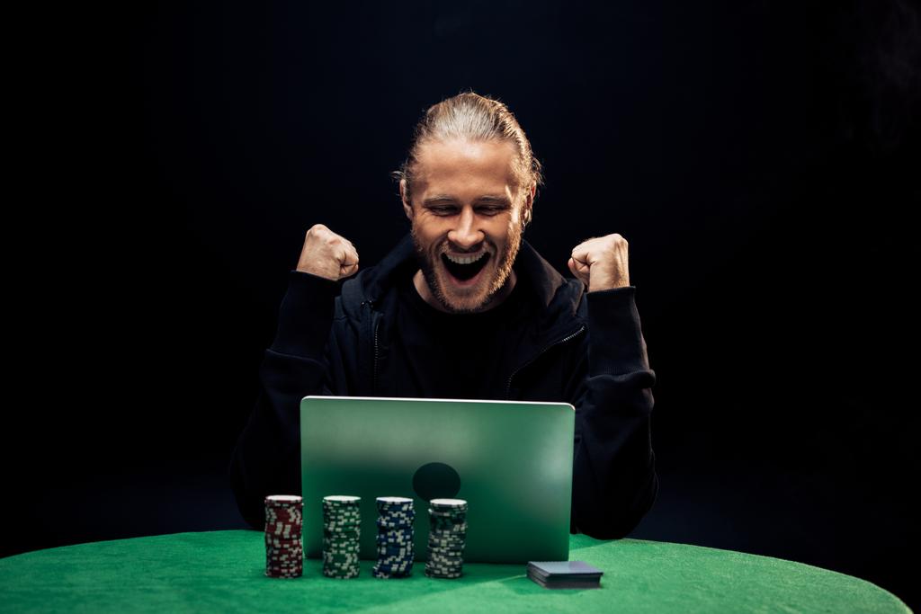 KYIV, UCRANIA - 20 de agosto de 2019: hombre feliz gesticulando mientras usa el ordenador portátil cerca de fichas de póquer aisladas en negro
  - Foto, Imagen