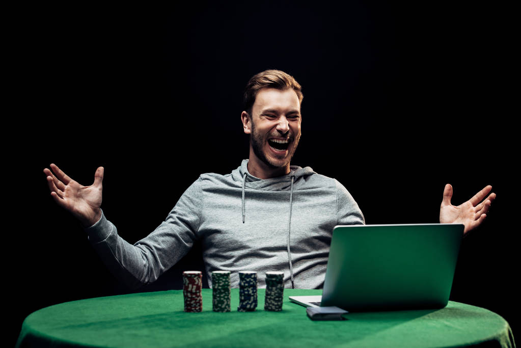 KYIV, UCRANIA - 20 de agosto de 2019: enfoque selectivo del hombre feliz gesticulando cerca de fichas de póquer cerca del ordenador portátil aislado en negro
  - Foto, imagen