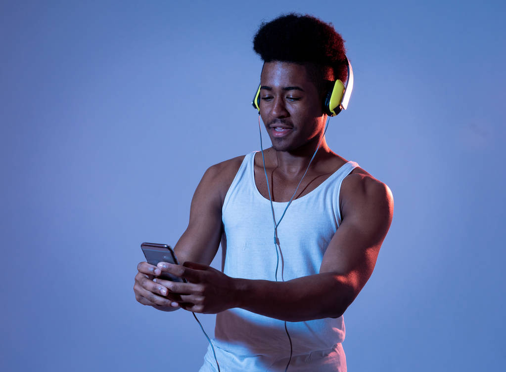 Silny African amerykański młody człowiek słuchając muzyki w smartfonie aplikacja szuka najlepszej piosenki online ze słuchawkami śpiew i taniec. Portret z disco Dance Floor światło w muzyce i technologii. - Zdjęcie, obraz