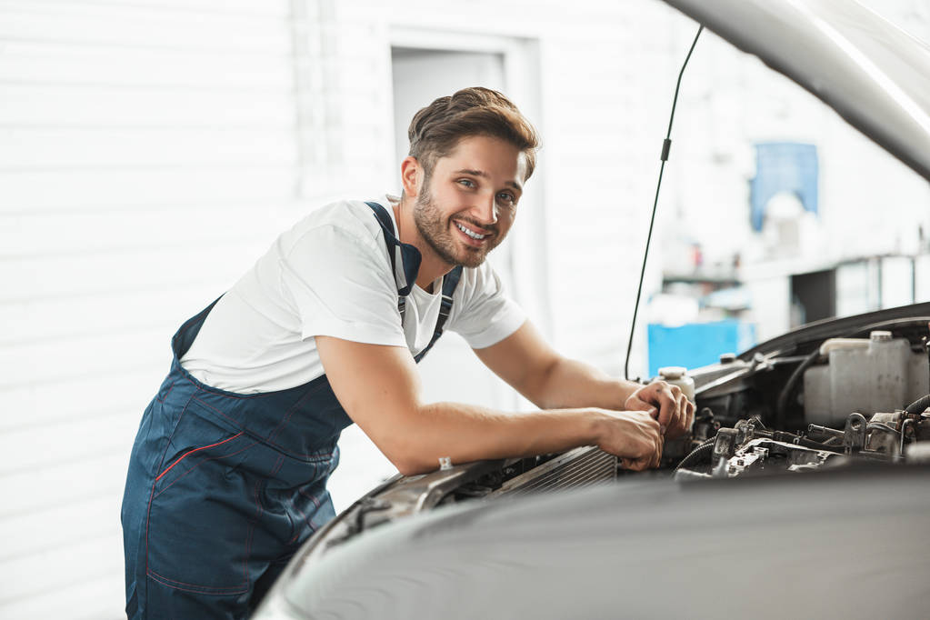 młody przystojny uśmiechnięty mechanik noszący mundur mocujący silnik w masce samochodu pracujący w dziale serwisowym - Zdjęcie, obraz