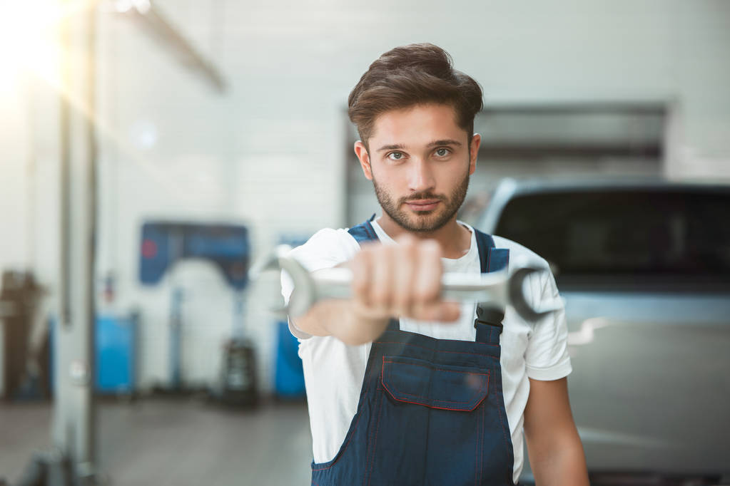 νεαρός όμορφος μηχανικός φορώντας στολή κρατώντας κλειδί στο χέρι του στο κέντρο εξυπηρέτησης αυτοκινήτων - Φωτογραφία, εικόνα