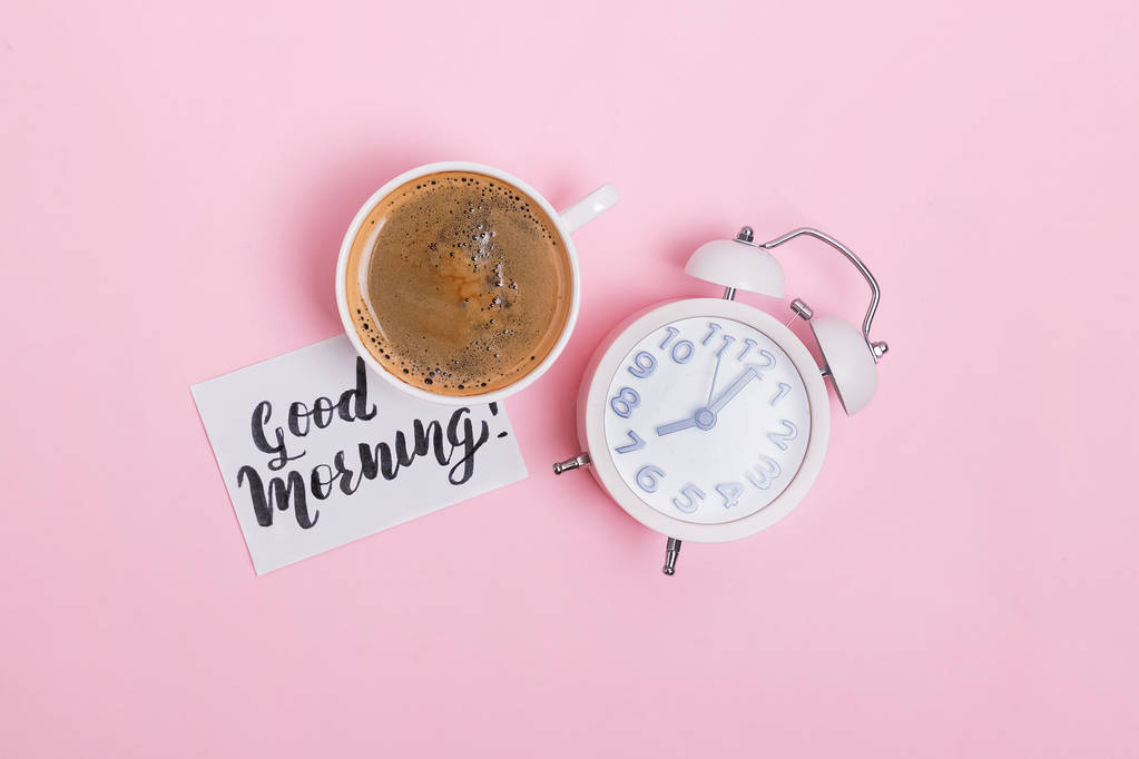 Analog alarm clock, cup of coffee and note with text good morning on pink background. - Photo, Image