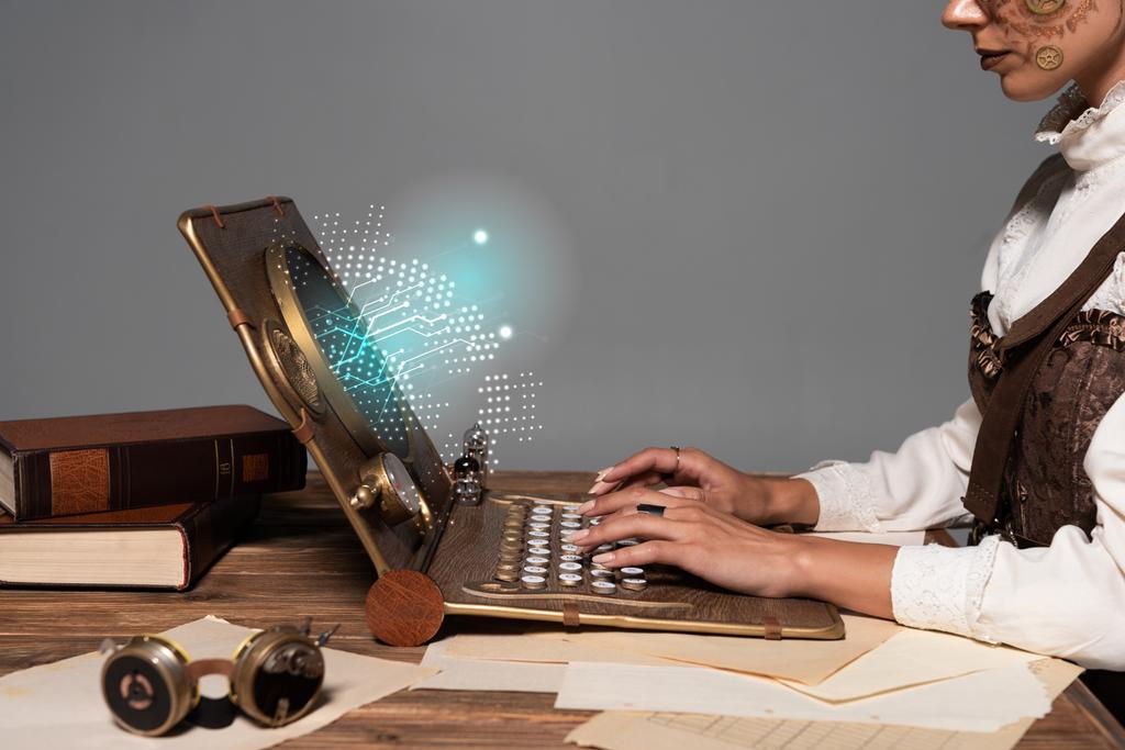 cropped view of woman typing on steampunk laptop with glowing digital illustration at table isolated on grey - 写真・画像