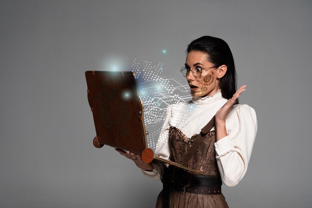 schockierte Steampunk-Frau in Brille mit Vintage-Laptop mit glühender digitaler Illustration isoliert auf grau - Foto, Bild