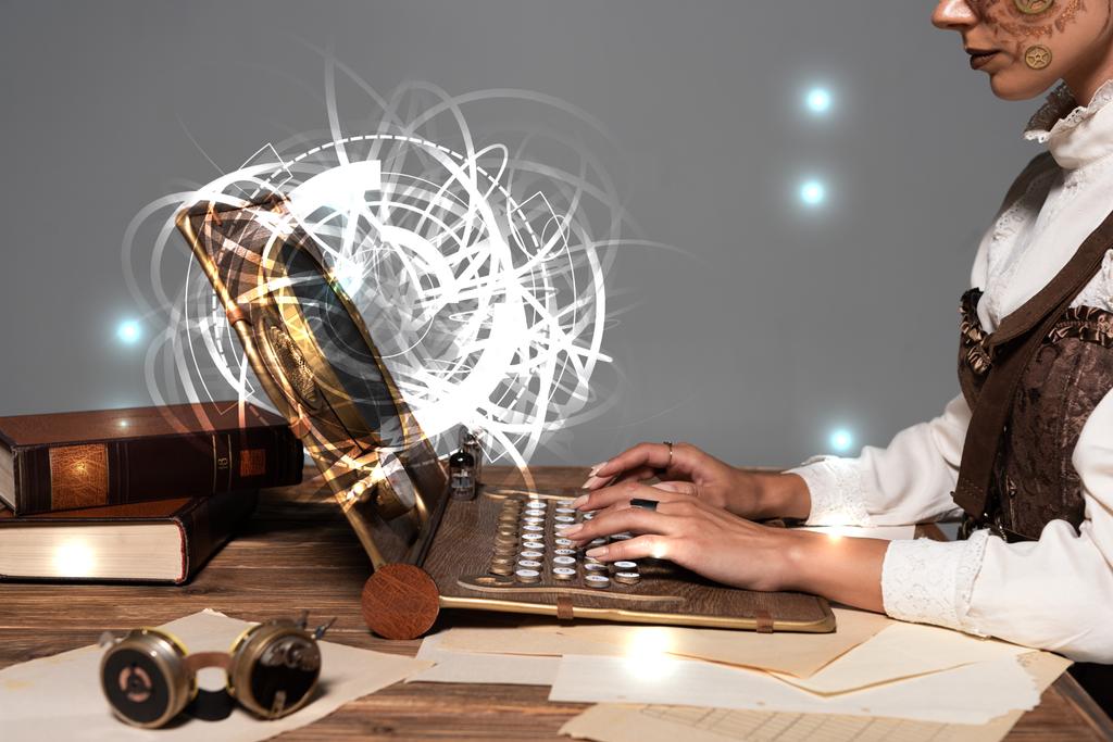 abgeschnittene Ansicht einer Frau beim Tippen auf einem Steampunk-Laptop mit glühender digitaler Illustration am Tisch isoliert auf grau - Foto, Bild