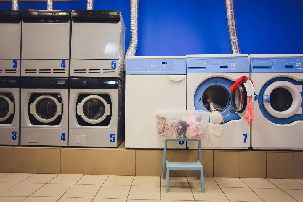 Profesjonalne pralki z koszykami, pełne ubrań w samoobsługowej pralni. - Zdjęcie, obraz