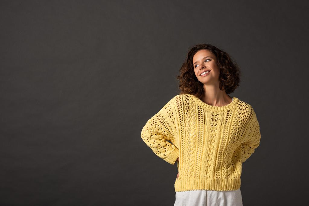 happy curly woman in yellow knitted sweater looking away on black background - Photo, Image