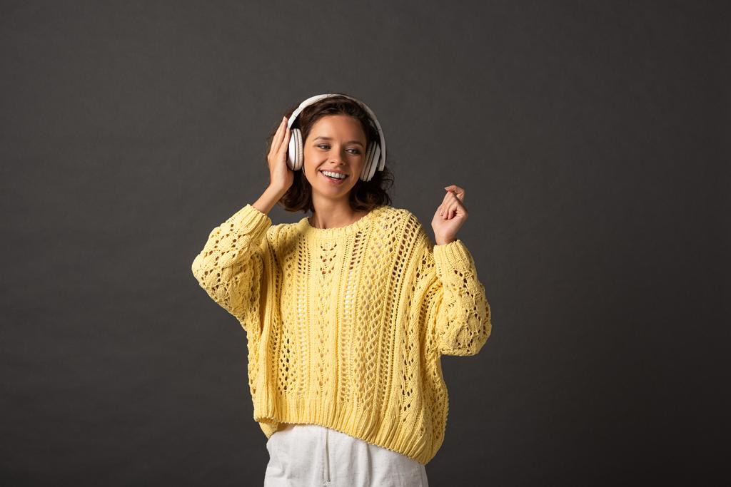 Lächelnde, lockige Frau in gelbem Strickpullover, die Musik über Kopfhörer auf schwarzem Hintergrund hört - Foto, Bild