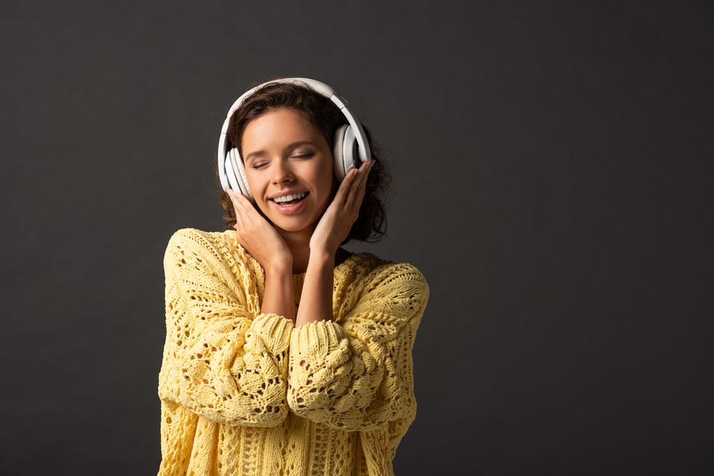 smiling curly woman with closed eyes in yellow knitted sweater listening music in headphones on black background - Photo, Image
