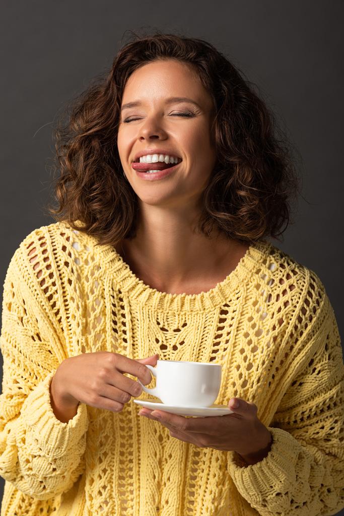 glückliche lockige Frau in gelbem Strickpullover, die Zunge mit geschlossenen Augen zeigt und eine Tasse Kaffee und Untertasse auf schwarzem Hintergrund hält - Foto, Bild