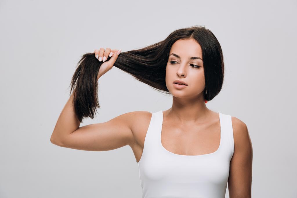 брюнетка красивая женщина глядя на длинные прямые здоровые и блестящие волосы изолированы на сером
 - Фото, изображение