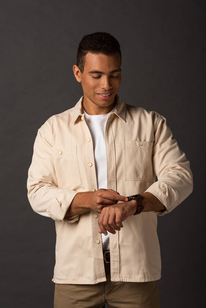 smiling handsome mixed race man in beige shirt looking at wristwatch on black background - Photo, Image