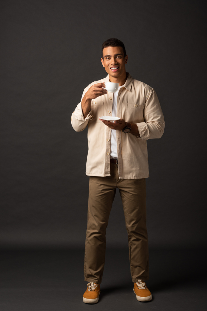 lächelnder gutaussehender Mischlingsmann in beigem Hemd mit Tasse Kaffee und Untertasse auf schwarzem Hintergrund - Foto, Bild